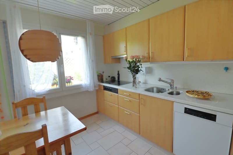 Bright kitchen with light wooden cabinets, white countertops, a sink, a dishwasher, a wooden dining table with chairs, and a vase of flowers on the counter.