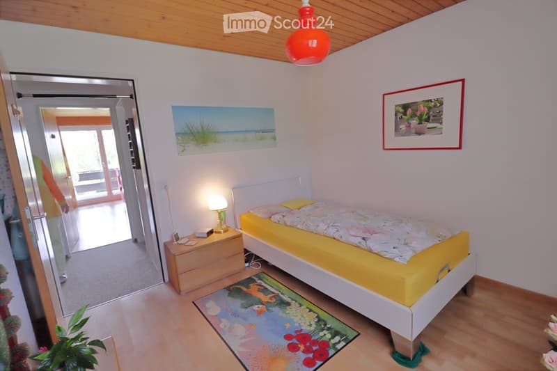 Cozy bedroom with a single bed, nightstand, lamp, and artwork on the walls. The room features wooden flooring and a colorful rug.