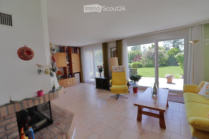Bright living room with a fireplace, yellow sofa, coffee table, armchair, bookshelf, and large windows overlooking a garden.