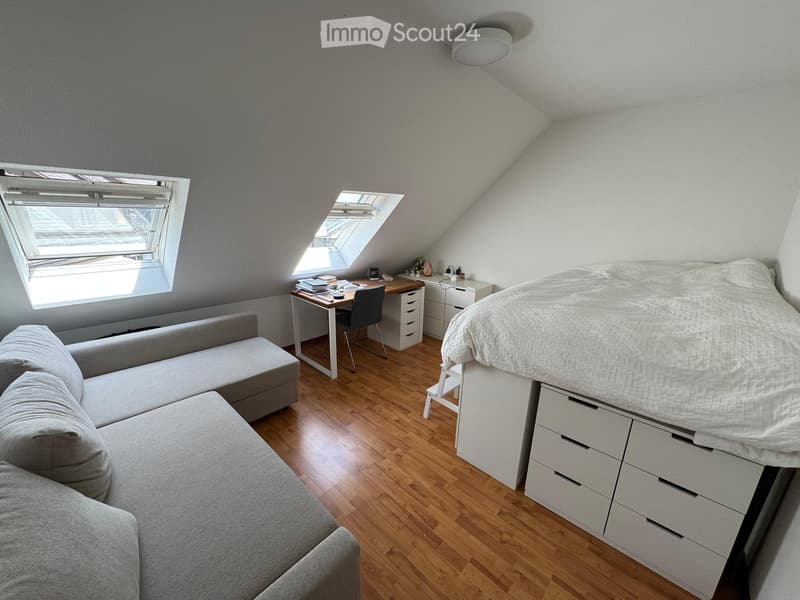 Cozy attic bedroom with wooden flooring, a raised bed with drawers, a sofa, a desk with a chair, and a nightstand.