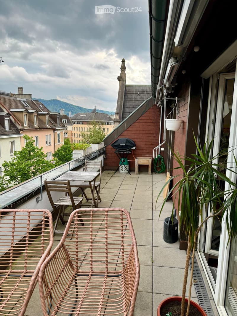Balcony with a table, chairs, a bench, a potted plant, and a grill, overlooking a cityscape with buildings and greenery.