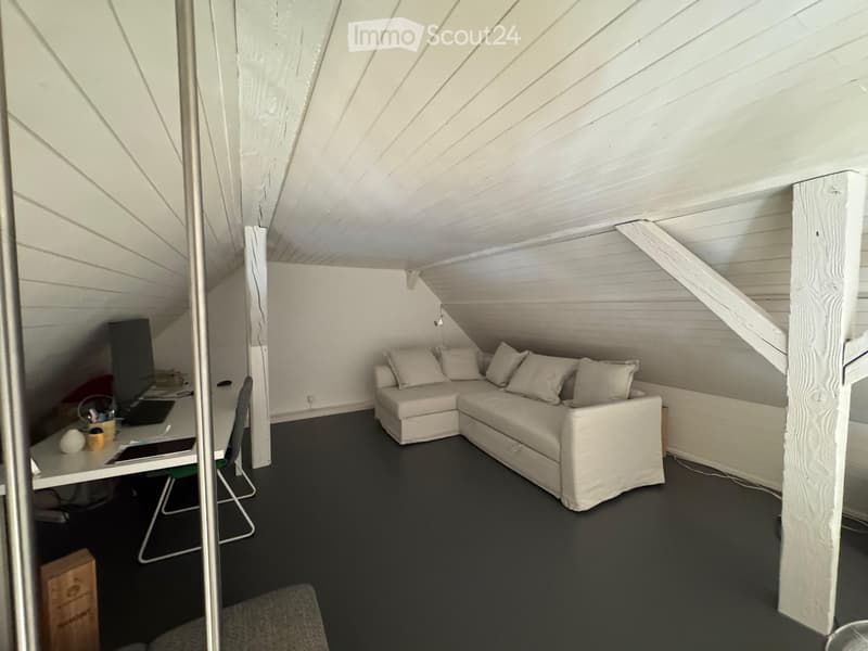 Loft-style room with slanted wooden ceiling, white walls, dark flooring, a white sectional sofa, a desk with a laptop, and a chair.