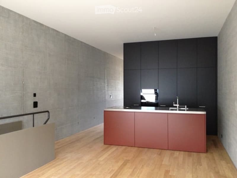 Modern kitchen with a red island, black cabinets, and a sleek faucet. The space features light wooden flooring and a concrete accent wall.