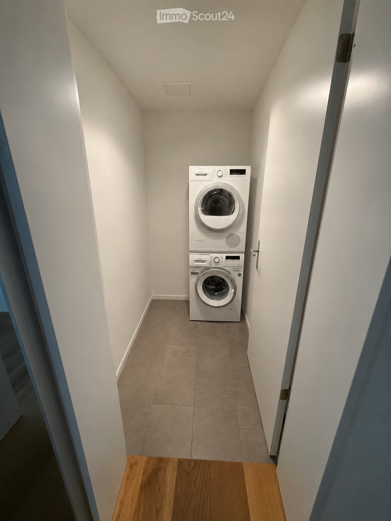 A narrow, well-lit room with a washing machine and dishwasher stacked vertically, beige tiled floor, white walls, and wooden flooring at the entrance.