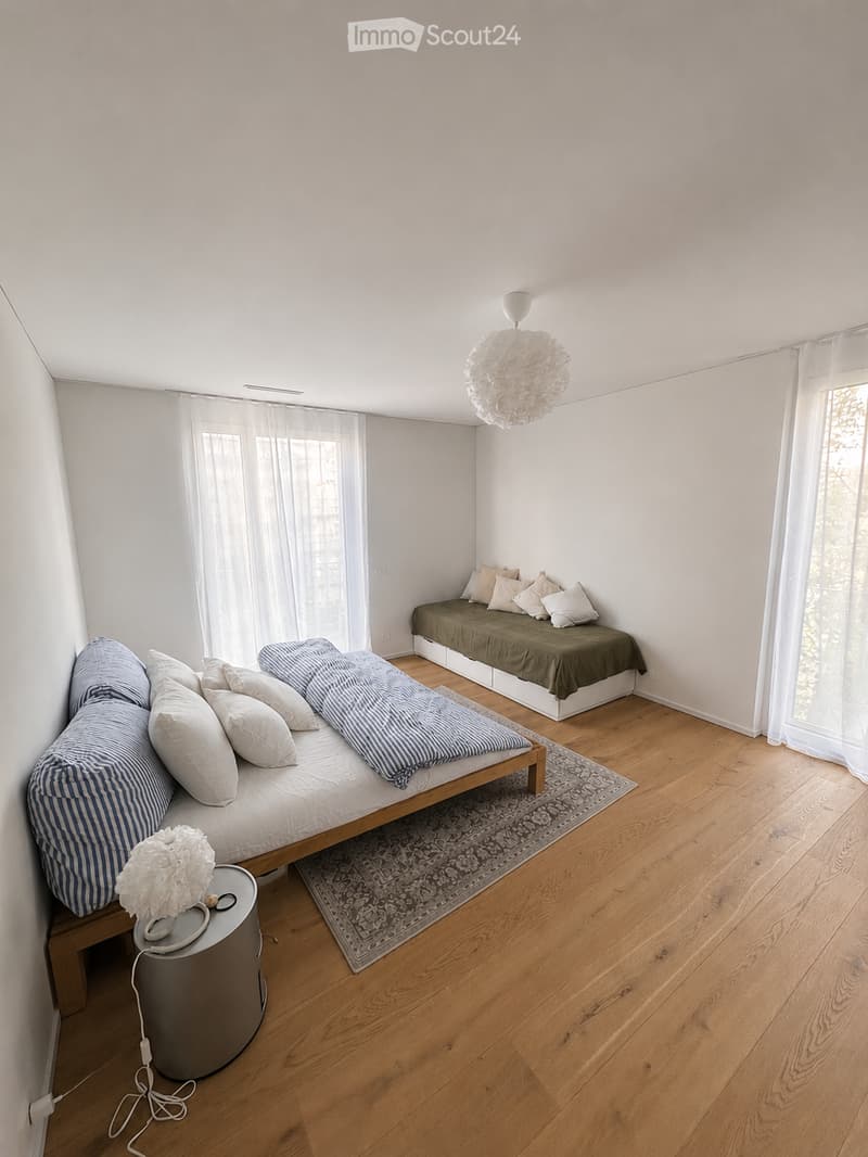 A modern, minimalist bedroom with wooden flooring, two beds (one with blue and white striped bedding, the other with green bedding), a small round table with a decorative item, and a patterned rug.