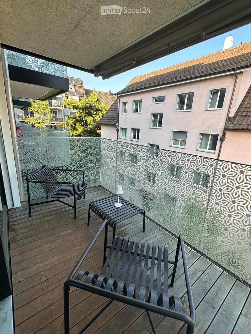 Balcony with wooden decking, black metal chairs, small table with a lamp, glass railing with leaf pattern, overlooking neighboring buildings and trees.