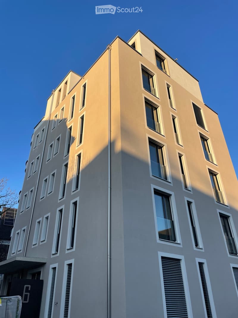 Modern multi-story building with light gray facade, white-framed windows, and a sleek design. The building features a mix of large windows and smaller rectangular windows, with a clean and contemporary architectural style.