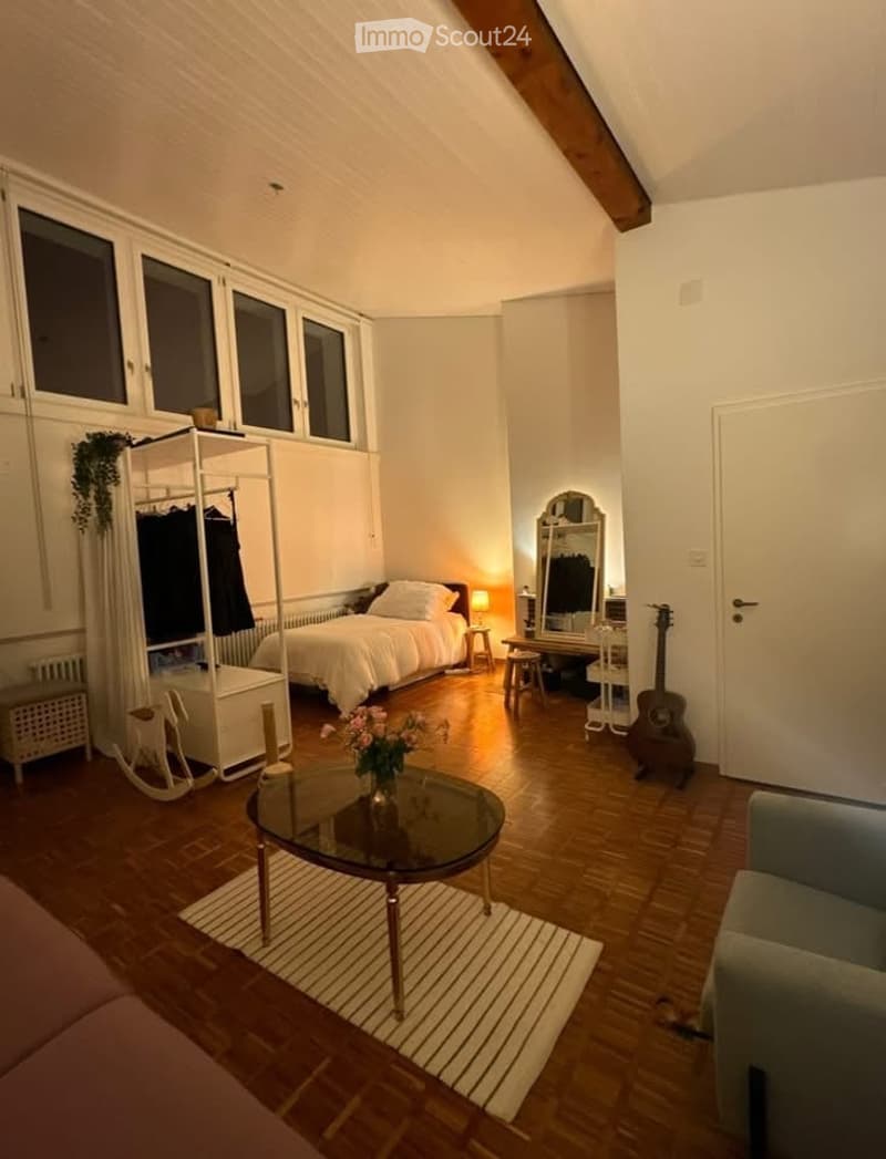 Cozy attic bedroom with wooden floors, large windows, a bed, a sofa, a coffee table, a guitar, a mirror, and a wardrobe.
