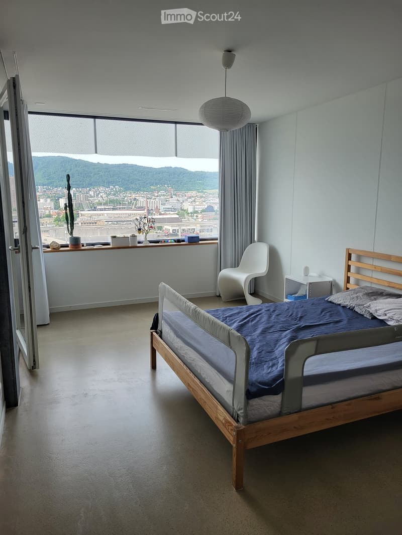 Modern bedroom with a wooden bed, blue bedding, white chair, nightstand, and a view of the city and mountains through large windows.