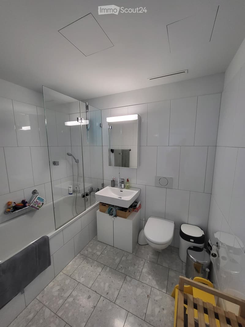 Modern bathroom with a bathtub, sink, toilet, mirror, and shower area. Features tiled walls and floor, a built-in storage cabinet under the sink, and a towel hanging on the side of the bathtub.