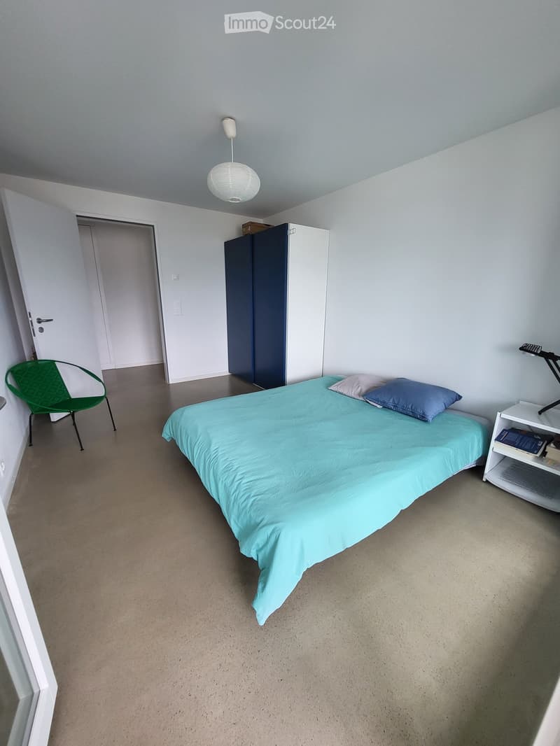 A modern, minimalist bedroom with a single bed, a green chair, a white wardrobe, a nightstand, and a hanging lamp.