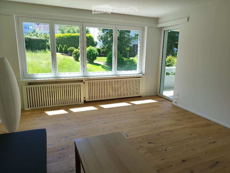 Empty room with wooden flooring, large windows, radiators, and a sliding glass door leading to a balcony.