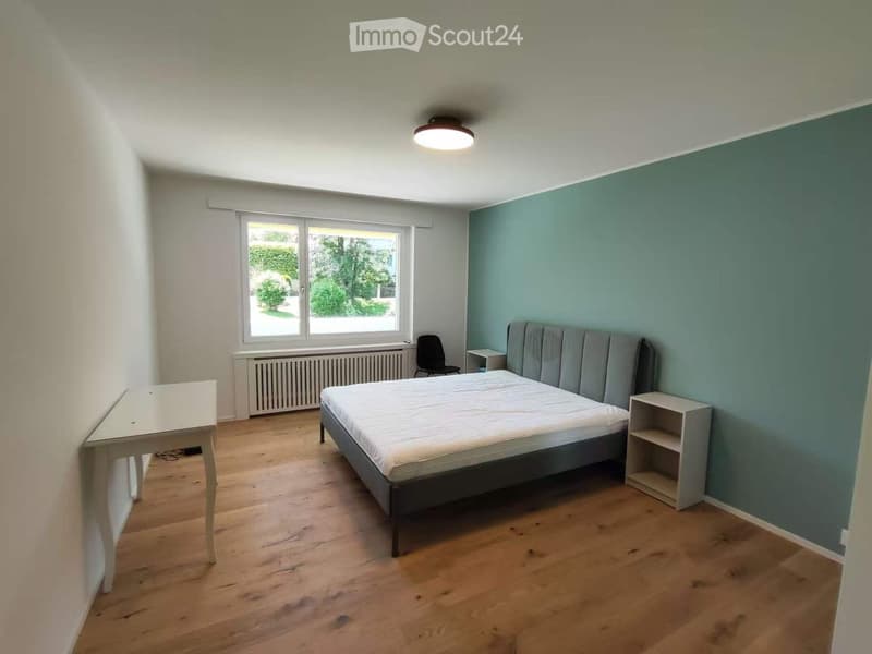 A furnished bedroom with a bed, a chair, a small table, and a shelf, featuring wooden flooring and a large window with a view of greenery.