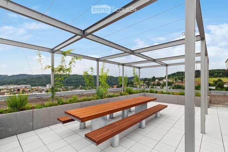 A modern, open-air terrace with a wooden table and benches, surrounded by greenery and offering a scenic view of the city and hills.
