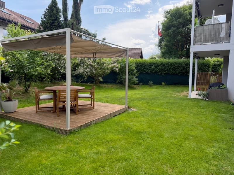 Garden with a wooden deck, umbrella, table, and chairs, surrounded by green grass and trees.