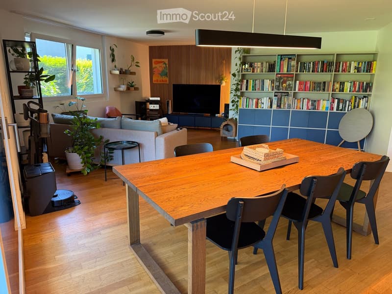 Open-plan living and dining area with a large wooden dining table, modern chairs, a sofa, bookshelf, TV, and plants.