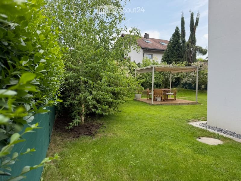 Well-maintained garden with a wooden patio, dining set, and a pergola, surrounded by lush greenery and trees.