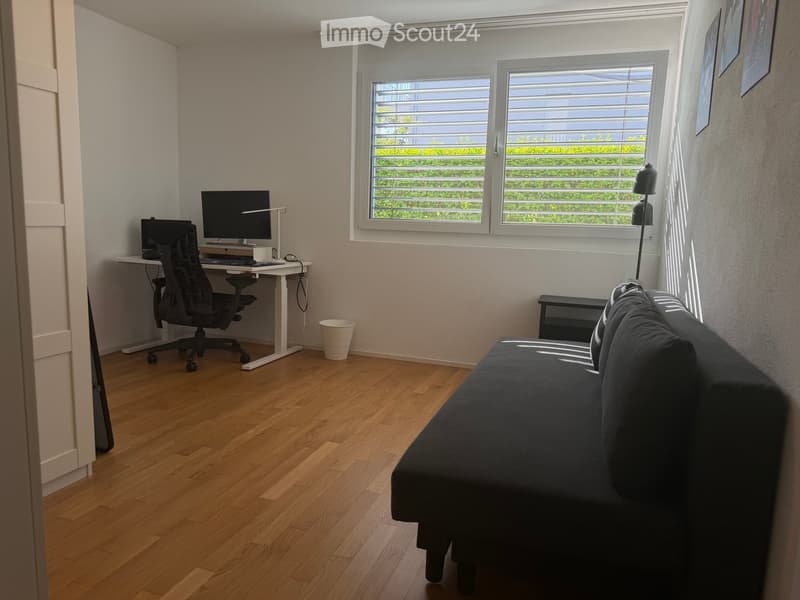 A modern, multi-functional room with a dark grey sofa, a white desk with a computer and chair, and large windows with blinds. The room features wooden flooring and a small trash bin near the desk.