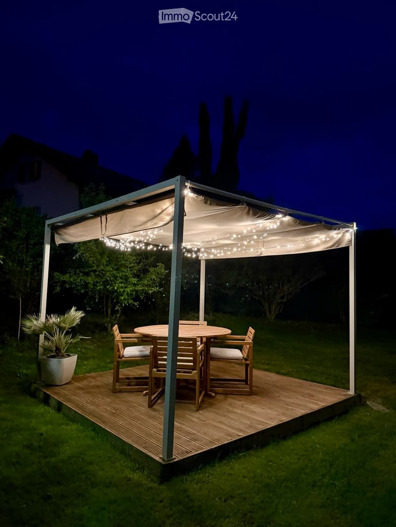 A wooden deck with a round table and four chairs, covered by a canopy with string lights, surrounded by greenery.