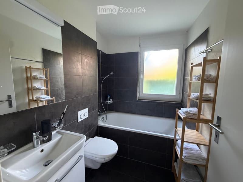 Modern bathroom with dark tiles, a large window, a bathtub, a toilet, a sink, and wooden towel racks holding towels.