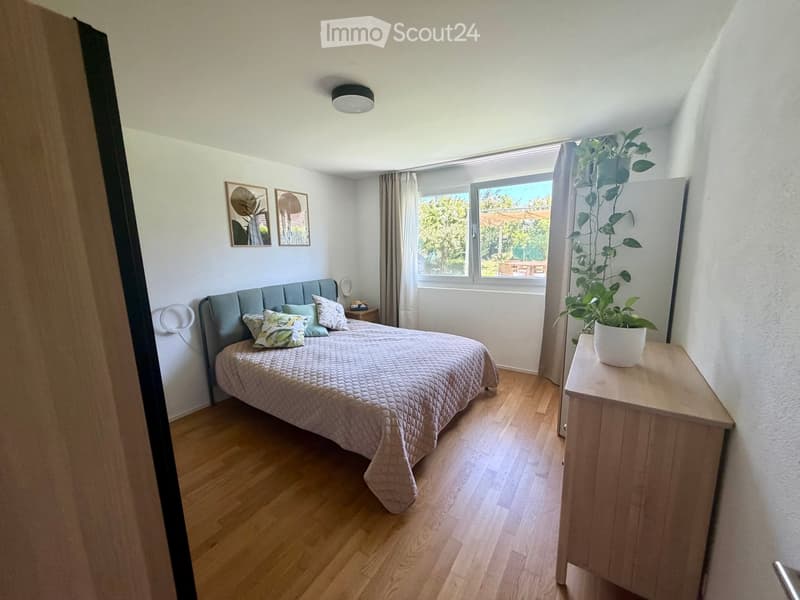 Cozy bedroom with wooden flooring, a large bed with pillows and a quilt, a nightstand with a plant, and two picture frames on the wall.