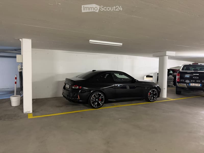 Underground parking area with two cars parked, one black and one dark blue, along with a white bucket and a white pillar.