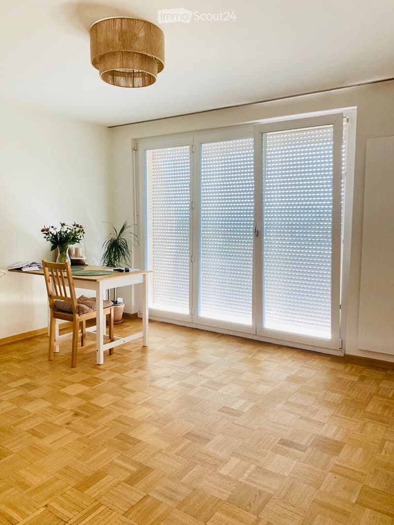 Bright room with wooden herringbone flooring, large sliding glass doors with blinds, a round wooden table with a chair, a vase with flowers, and a hanging woven lamp.