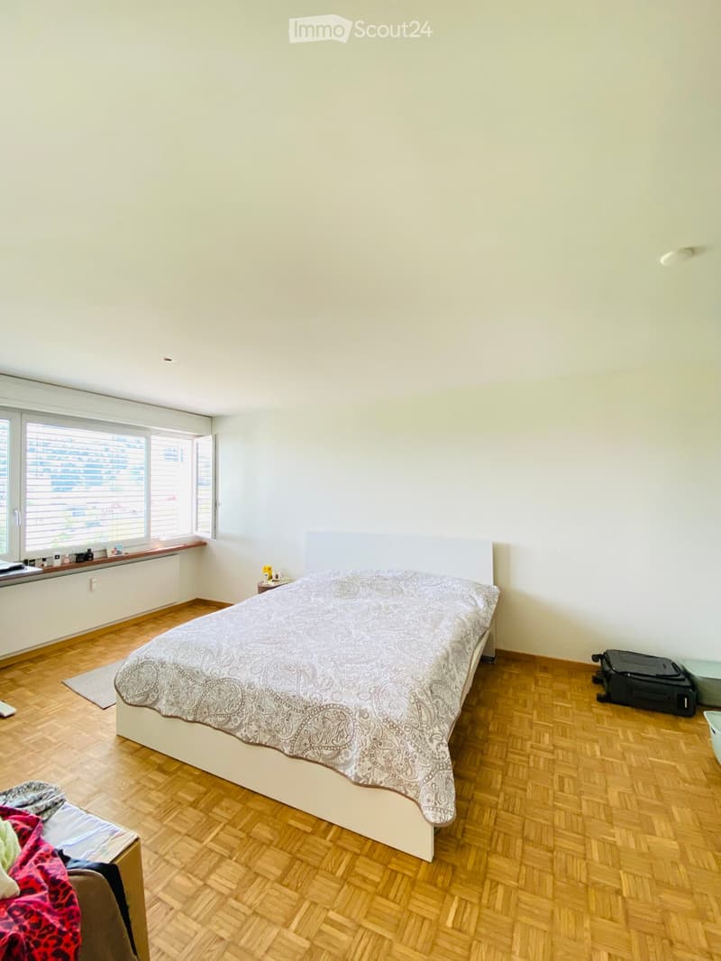Bright bedroom with wooden flooring, large window with blinds, bed with patterned bedspread, nightstand, and a suitcase on the floor.