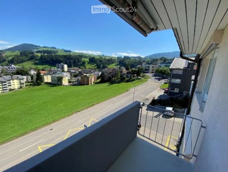 Balcony with a scenic view of a town, green fields, and mountains under a clear blue sky.