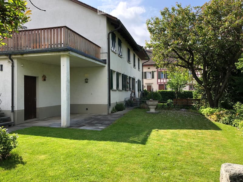 Well-maintained garden with green lawn, stone pathway, and a small fountain. The house features a balcony and a covered entrance area.