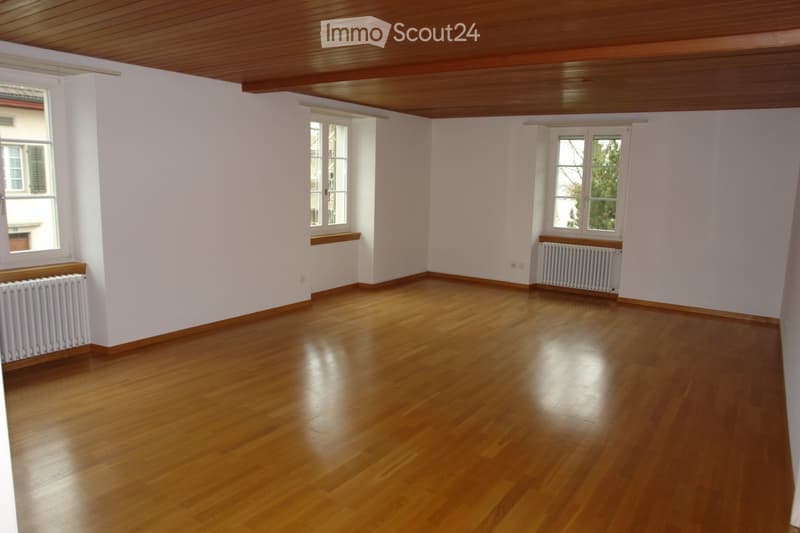 Empty room with wooden flooring, white walls, and multiple windows with radiators beneath them. The ceiling has wooden paneling.