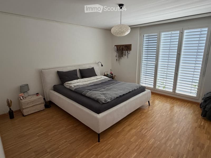 Modern bedroom with a large bed, nightstand, lamp, and wall decor. Large windows with blinds and wooden flooring.