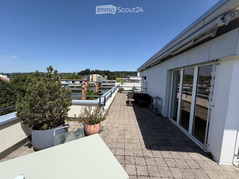 Balcony with tiled floor, white table, chairs, potted plants, and a view of the city and greenery.