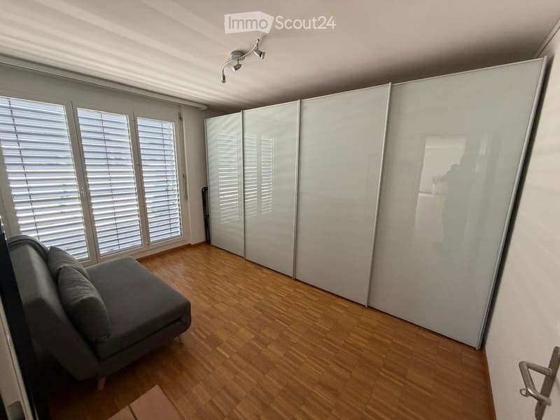 Modern living room with wooden flooring, large windows with blinds, and a grey sofa.