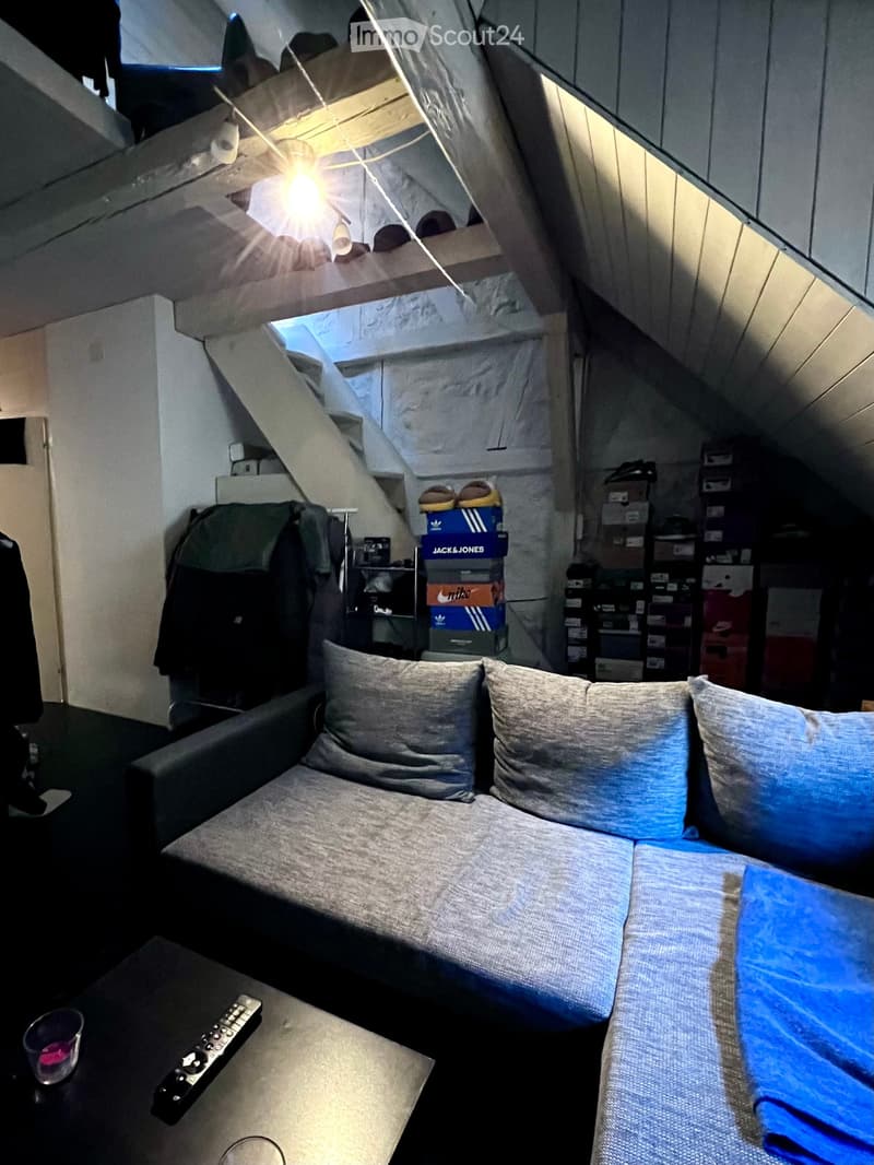 Cozy attic room with a grey sofa, coffee table, remote control, and cups. The room features exposed wooden beams and a skylight.