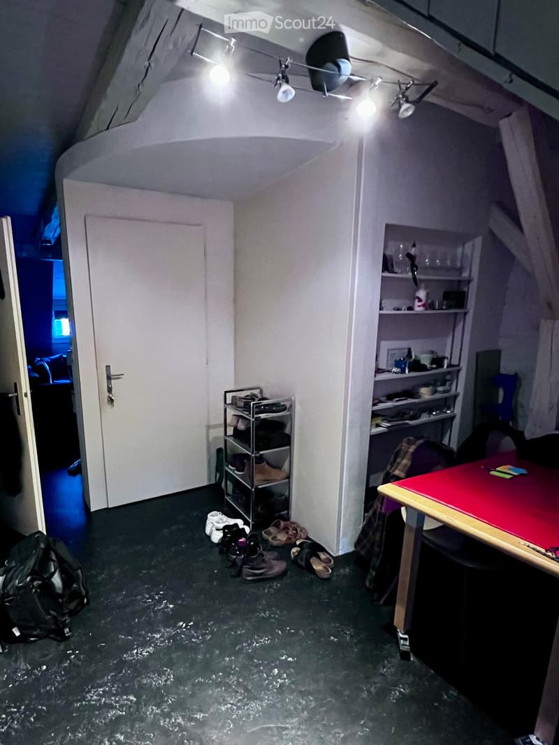 Small attic room with slanted ceilings, a table with a red surface, a chair, a shelf with various items, and a backpack on the floor.