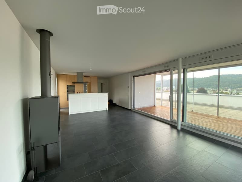 Open-plan living and kitchen area with dark tiled floor, white kitchen island, wooden deck, and large windows offering a scenic view.