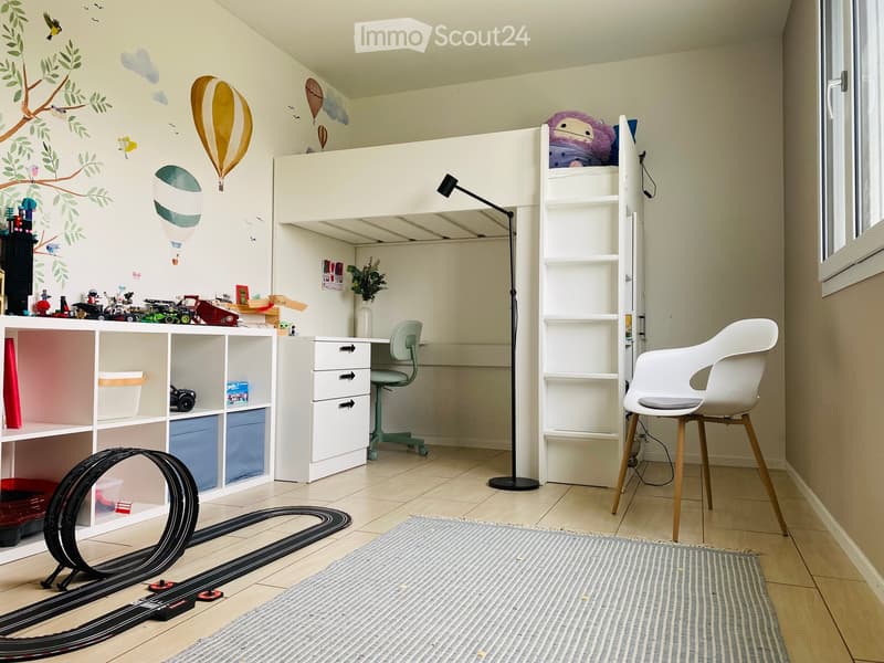 A well-lit children's bedroom with a lofted bed, desk, shelves, and a toy car track on the floor.