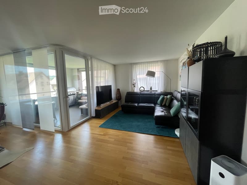 Modern living room with wooden flooring, large glass sliding doors leading to a balcony, a black leather sofa, a coffee table, a television, a floor lamp, and a green rug.
