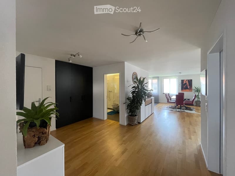 Bright hallway with wooden flooring, leading to a dining area with chairs and a table, and a bathroom with a toilet and shower. Potted plants are placed along the walls.