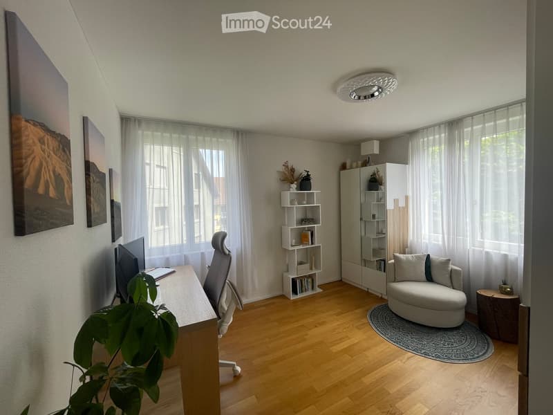 A modern, well-lit room with wooden flooring, large windows, a desk with a screen, a chair, a round sofa, a shelf, and a plant. The room features contemporary decor with artwork on the walls and a sleek ceiling light.
