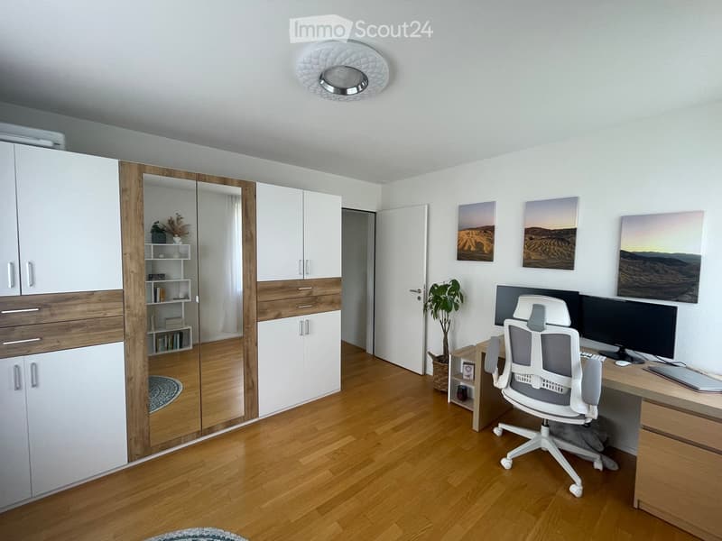 Modern, well-lit room with wooden flooring, white cabinetry, a desk with a computer and chair, and decorative artwork on the walls.