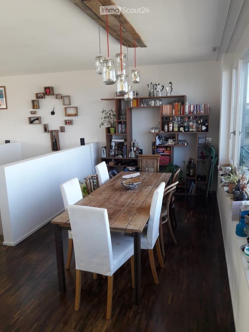 Dining area with a wooden dining table, white chairs, shelves with books and bottles, and pendant lights.