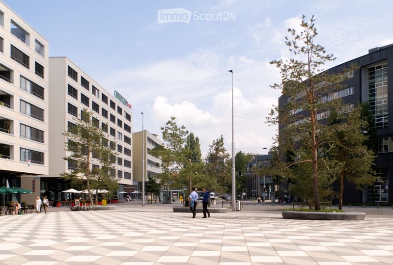 A spacious urban plaza with a checkered pavement, surrounded by modern buildings, trees, and outdoor seating areas with umbrellas. People are walking and sitting, enjoying the area.