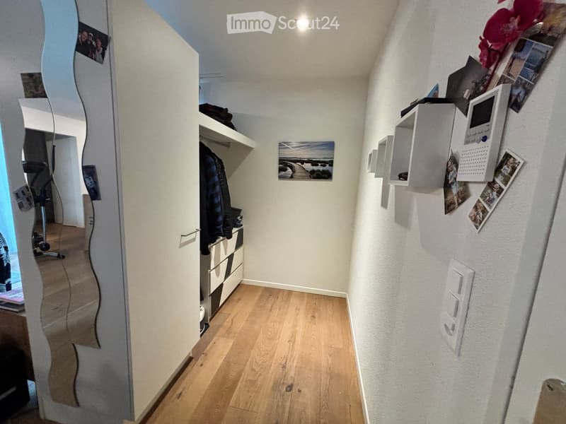 A narrow hallway with light wooden flooring, white walls, and built-in shelving units. Photos and a calendar are displayed on the wall, and a small closet with clothes is visible.
