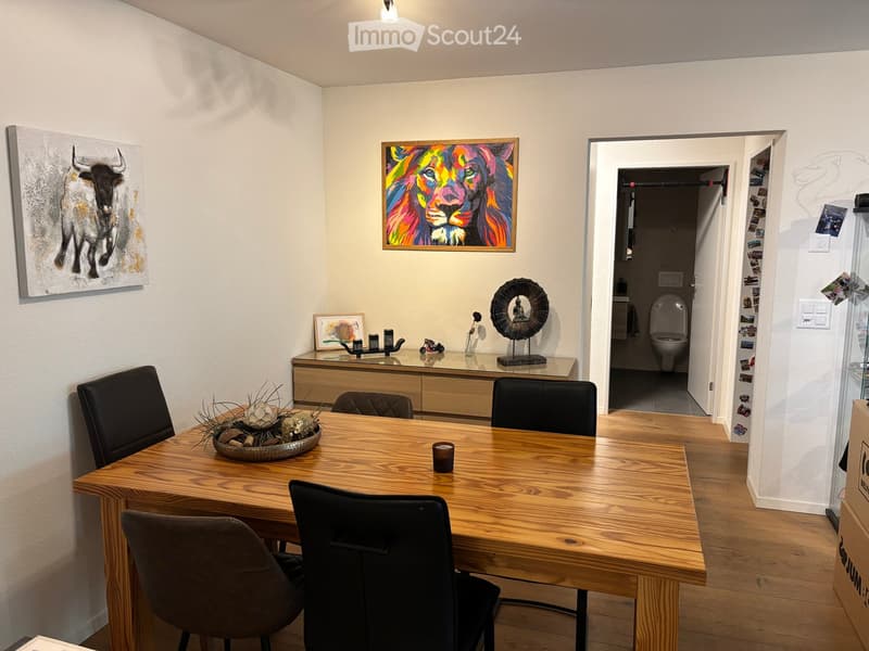 Dining room with wooden table, chairs, cabinet, decorative items, and artwork on walls. Open doorway leads to a bathroom.
