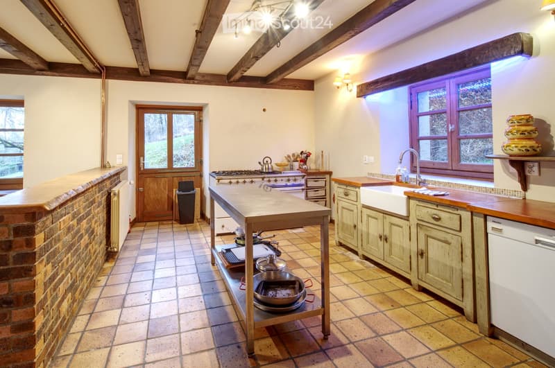 Bright, rustic kitchen with wooden beams, exposed brick wall, and large windows. Features a central island, stove, sink, and various kitchen appliances.