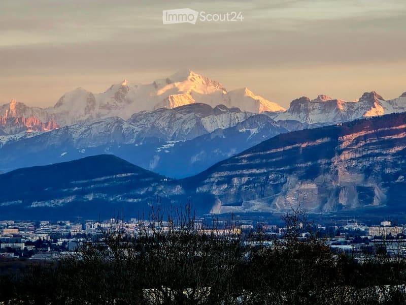 Panoramic view of snow-capped mountains and a cityscape in the foreground, with a warm sunset lighting the peaks.