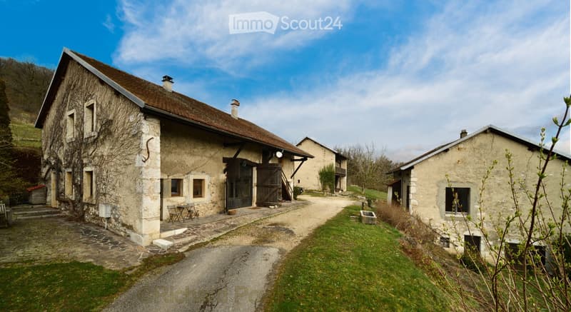 A rustic stone house with a brown tiled roof, multiple chimneys, and a wooden entrance door. The house is surrounded by a gravel driveway, grassy areas, and smaller outbuildings.