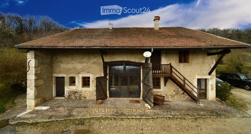 Two-story stone house with a tiled roof, wooden doors, and a balcony accessible via wooden stairs. The house features a stone-paved courtyard with a small table and chairs, and a parked car is visible nearby.